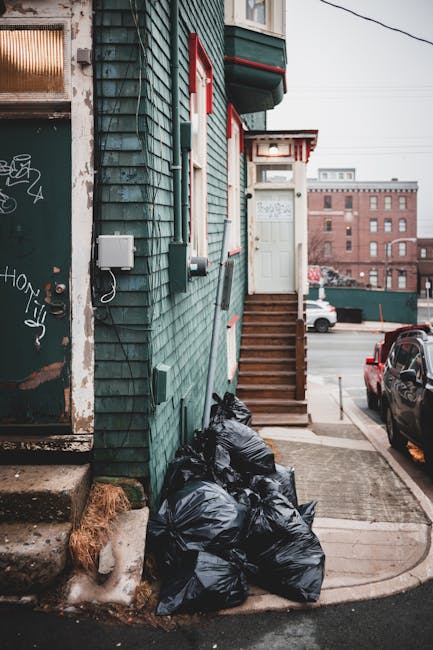 A scene outside a building with green wooden exterior siding featuring red and white window frames and a set of wooden stairs leading to a white door. In the foreground, there are several black garbage bags piled against the building’s lower wall on the concrete pavement. The bags appear full and slightly shiny, indicating they are filled with waste. To the left, there's a weathered metal door with graffiti and paint marks, with a small step leading up to it. A portion of a small window with a metal grid is also visible. In the background, a cityscape with brick buildings, parked cars, and an overcast sky can be seen, suggesting an urban environment. The scene is lit with natural diffuse daylight, emphasizing the textures of the wooden siding, the garbage bags, and the surrounding structures, reflecting common arrangements during private rubbish clearance or alternative waste disposal in a residential or commercial area managed by independent waste collection services like rubbishclearancefinsburypark.co.uk.
