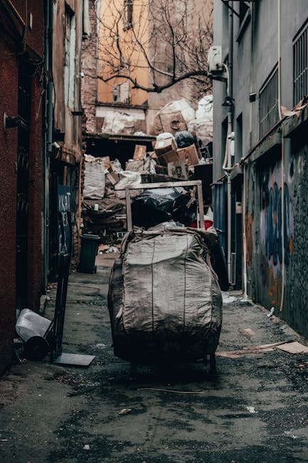 A scene outside a building with green wooden exterior siding featuring red and white window frames and a set of wooden stairs leading to a white door. In the foreground, there are several black garbage bags piled against the building’s lower wall on the concrete pavement. The bags appear full and slightly shiny, indicating they are filled with waste. To the left, there's a weathered metal door with graffiti and paint marks, with a small step leading up to it. A portion of a small window with a metal grid is also visible. In the background, a cityscape with brick buildings, parked cars, and an overcast sky can be seen, suggesting an urban environment. The scene is lit with natural diffuse daylight, emphasizing the textures of the wooden siding, the garbage bags, and the surrounding structures, reflecting common arrangements during private rubbish clearance or alternative waste disposal in a residential or commercial area managed by independent waste collection services like rubbishclearancefinsburypark.co.uk.