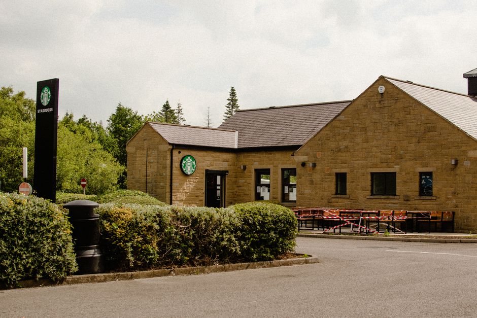 A single-story commercial building constructed from light brown stone bricks, featuring a sloped dark gray tiled roof, is situated on a paved area adjacent to a road. The building's facade includes two small, rectangular black-framed windows and a central entrance door with a glass panel. Above each window and door, there are small security or sensor lights attached to the wall. To the left of the building, a tall, vertical black sign with the Starbucks logo, featuring a green mermaid emblem on a white background, is prominently displayed. In front of the building, dense, well-maintained green shrubs form a hedge along the sidewalk, partially obscuring a black cylindrical waste bin placed within the greenery. Outside, the public space is free of litter, indicating regular maintenance. In the background, a line of tall, slender trees and a partly cloudy sky create a natural backdrop, with no other structures immediately visible. The scene suggests an urban or suburban setting where private waste management, possibly managed by [COMPANY_NAME], handles rubbish removal from commercial premises like cafes or shops, contributing to the tidy appearance of the area.