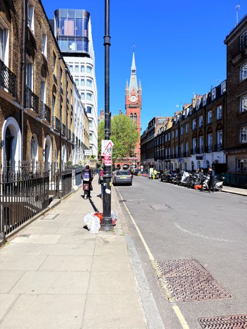 A wide urban street scene showing a clear, sunny day with bright blue skies. On the left side, there are Victorian-style brick terraced buildings with arched windows and black iron railings. A sidewalk runs alongside these buildings, with two individuals walking and carrying materials, while a large white plastic bag filled with rubbish is placed on the pavement near a lamppost. The lamppost is centrally positioned in the image, with a pedestrian crossing sign attached to it. On the right side, several parked motorcycles and bikes are lined up along the curb, adjacent to more terraced buildings similar in style to those on the left. In the background, a tall, historic clock tower with a pointed steeple and a clock face dominates the scene, set against the blue sky. The street appears to be in a residential or mixed-use area, and the image subtly illustrates the context of local or private waste collection, with the rubbish bags and litter possibly prepared for collection by a private waste management service like Rubbish Clearance Finsbury Park.
