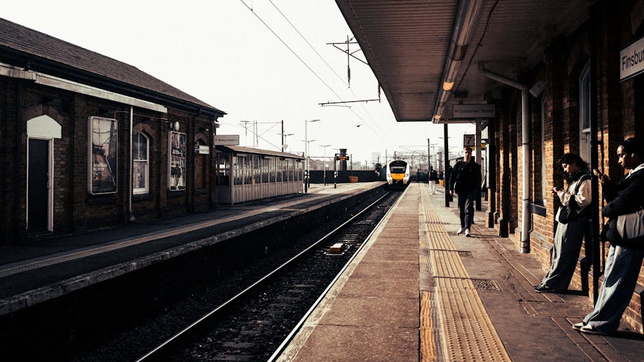At Finsbury Park station platform, a yellow and white train approaches in the distance, with several passengers waiting along the edge of the brick platform. The station building on the right features a brick facade, large windows, and a sheltered canopy extending over the waiting area, supported by metal posts. Some passengers are standing casually, wearing jackets and casual clothing, with one person looking at their phone and another with arms crossed. A tactile paving strip runs along the platform’s edge, indicating safe distance from the tracks. In the background, railway tracks extend into the distance, with overhead electrical wires and signals visible under a partly cloudy sky. The scene is lit with natural daylight, creating soft shadows and highlighting the textured surfaces of the brick building and platform. While this image primarily captures a busy station environment, the clear view of the tracks and station infrastructure emphasizes the importance of efficient passenger flow and safety, elements that are supported by independent or private forms of transportation and public transit management.