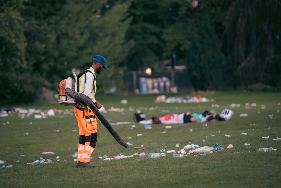 A man wearing high-visibility orange trousers, a white jacket with reflective strips, and a blue cap is using a leaf blower with a long black hose on a grassy park area. The man is cleaning up scattered litter, including plastic bottles, cups, and paper, which are spread across the ground in front of him. In the background, a person is lying on the grass near a large tree, and there are additional waste items and recycling bins visible further back, indicating a recent outdoor gathering or event. The scene is set outdoors with natural lighting, surrounded by trees and greenery, and the environment appears to be in need of waste management or cleaning. The image relates to the context of alternative waste handling, possibly emphasizing private rubbish clearance services such as those offered by rubbishclearancefinsburypark.co.uk, in contrast to municipal collection.