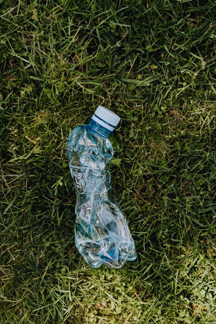 A clear plastic PET water bottle with a white cap lies discarded on a patch of lush green grass, with blades of grass partially covering the bottom of the bottle. The bottle appears crumpled and empty, with visible creases and folds in the plastic material, which has a shiny, reflective surface. The grass around the bottle is a mixture of fine and coarse blades, varying in shades of green, creating a natural environment that highlights the importance of responsible waste disposal. The scene suggests an outdoor setting, possibly a park or garden area, where littering might occur, and underscores the relevance of private rubbish removal services such as those provided by companies like rubbishclearancefinsburypark.co.uk. No other objects or environmental elements are visible in the scene, emphasizing the solitary piece of litter in a natural landscape.