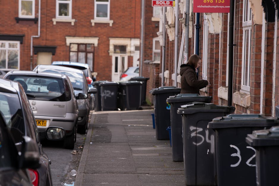 A row of large, dark grey plastic wheelie bins with white painted numbers on their sides are lined up along the pavement outside red-brick residential buildings. One person in a brown jacket is standing next to a bin, appearing to be opening or closing its lid. The bins are positioned in front of windows and doorways, with some bins slightly open or closed, indicating recent use. Several parked cars, mostly silver or grey, are aligned along the street, occupying parking spaces directly adjacent to the bins. The pavement is made of concrete slabs, and small bits of litter are scattered on the ground near the curb. In the background, additional residential brick buildings and more parked vehicles are visible, with typical urban street scene elements including a red 'Sold' sign attached to one of the buildings. The scene suggests a residential area with on-street waste collection or potentially private rubbish removal services, as evidenced by the presence of multiple bins designated for waste disposal, possibly coordinated by a local waste management company such as Rubbish Clearance Finsbury Park.
