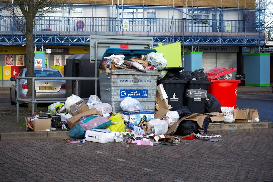 A collection of overflowing waste bins and surrounding rubbish on a paved street surface in an urban area. The waste bins include a large grey mixed paper and card bin, a black bin for general waste, and a bright red bin, all positioned on the sidewalk near a metal railings. The grey bin is open, revealing a disorganized pile of paper, cardboard, and packaging materials spilling out onto the ground. Additional trash, including cardboard boxes, plastic bags, paper packaging, and discarded items, are scattered around the bins. To the left, a parked silver car is visible, partially obscured by the waste. In the background, a building with a blue scaffolding structure and signage indicative of commercial premises can be seen, with the ground floor featuring storefronts with window displays. The scene is lit by natural daylight, emphasizing the clutter and the state of the rubbish, illustrating an example of private waste accumulation awaiting rubbish removal services, as offered by companies specializing in on-site clearance and independent waste disposal solutions in the area.